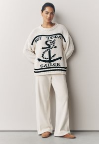 Jersey blanco de punto con diseño de ancla negra y texto "NO HOY, MARINERO". Combinado con pantalones anchos color crema y zapatos slip-on marrón claro.