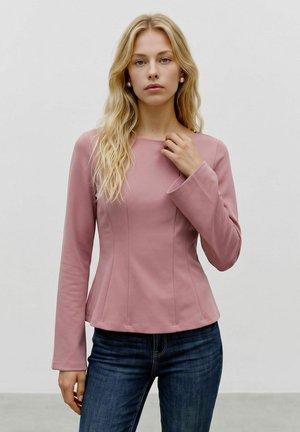 Femme blonde aux cheveux ondulés lâches, portant un haut rose moulant à manches longues et un jean bleu foncé, debout devant un fond clair uni.