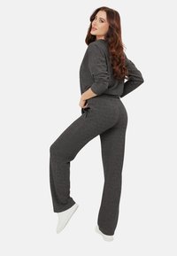 Kobieta z długimi brązowymi włosami, ubrana w ciemnoszary sweter dziany i spodnie, pozująca z uniesioną jedną nogą i dłońmi na biodrach, w białych trampkach.