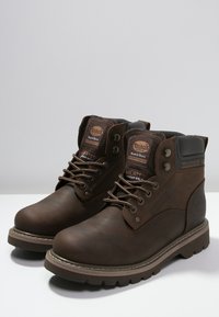 Botas aislantes marrones con parte superior de cuero, punta redonda, diseño de cordones, collar acolchado y suela de goma. Presenta detalles del logo en la lengüeta.