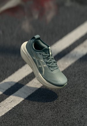 Scarpa da corsa grigia con suola bianca e logo del marchio, sospesa in aria sopra una pista di asfalto con linee bianche di corsia.