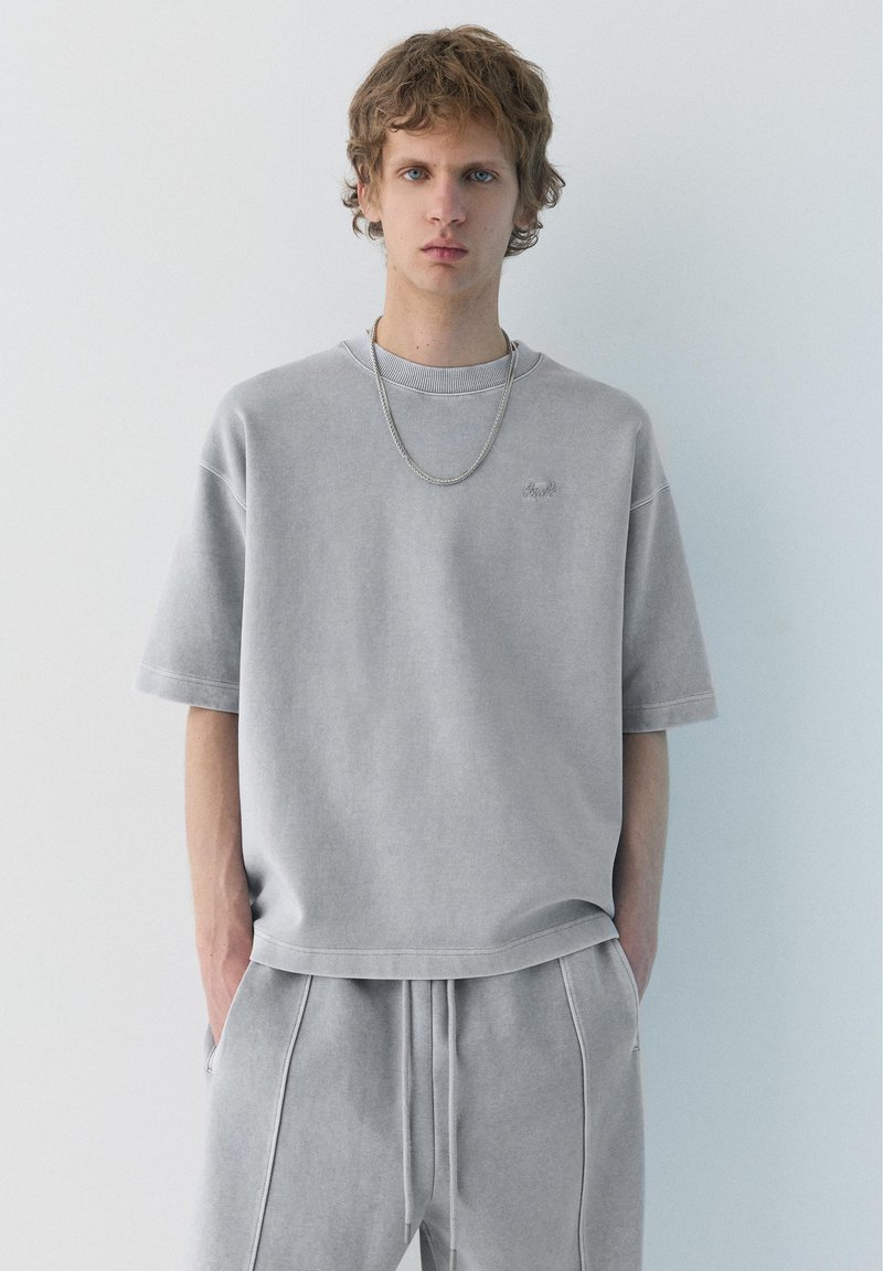 Jeune homme aux cheveux bouclés portant un t-shirt oversize gris clair et un pantalon de jogging assorti, debout les mains dans les poches devant un fond uni.