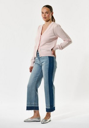 Sieviete, kas stāv, valkājot gaiši rozā kardiganu, divu toņu plaša piegriezuma džinsus un sudraba dzīvokļzābakus uz vienkārša balta fona.