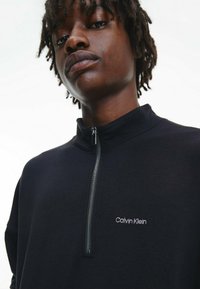 Sudadera negra con cremallera hecha de tela suave, con un cuello alto y el logotipo bordado de "Calvin Klein" en el lado izquierdo del pecho.