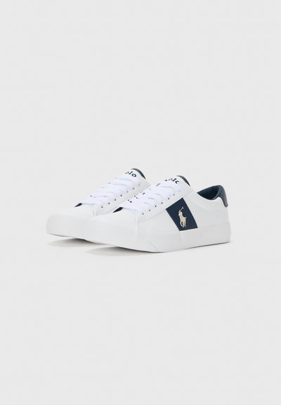 Baskets blanches basses avec des accents navy, des lacets et un logo d'un joueur de polo sur le côté, présentées sur un fond blanc uni.