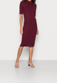 Abito bodycon borgogna con collo alto, maniche corte e texture liscia, abbinato a sandali trasparenti con tacco a blocco.