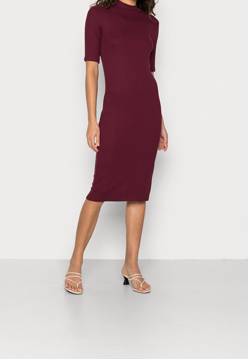 Abito bodycon borgogna con collo alto, maniche corte e texture liscia, abbinato a sandali trasparenti con tacco a blocco.