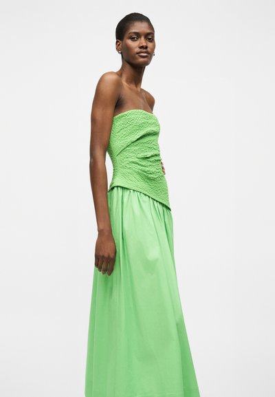 Γυναίκα φοράει ένα πράσινο τοπ χωρίς τιράντες με υφή και μια μακριά, ρέουσα πράσινη φούστα, στέκεται μπροστά σε ένα απλό λευκό φόντο.