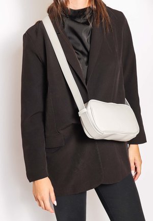 Personne portant un blazer noir et un pantalon avec un sac bandoulière blanc porté sur l'épaule, sur un fond uni.