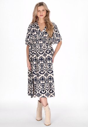 Kobieta stojąca, ubrana w czarno-białą wzorzystą sukienkę midi z krótkimi rękawami oraz beżowe botki, na jednolitym białym tle.