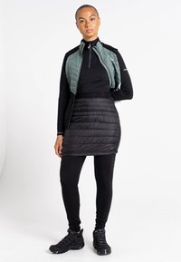 Schwarzer Pufferrock über schwarzen Leggings, kombiniert mit einer grünen, gesteppte, mit Reißverschluss versehenen Weste und einem schwarzen Langarmoberteil. Texturierte Materialien sichtbar.