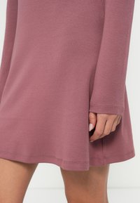 Φόρεμα με μακριά μανίκια σε αποχρώσεις του μωβ με απαλή υφή. Το τελείωμα φουσκώνει ελαφρώς, με ραμμένες λεπτομέρειες κατά μήκος του κάτω μέρους και στις άκρες των μανικιών.