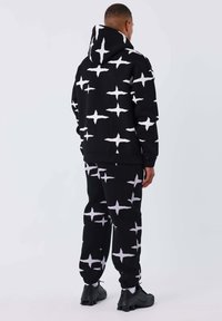 Conjunto de sudadera con capucha negra y pantalones deportivos que presenta un atrevido patrón de cruces blancas, con un ajuste holgado y una tela texturizada, diseñado para la comodidad.