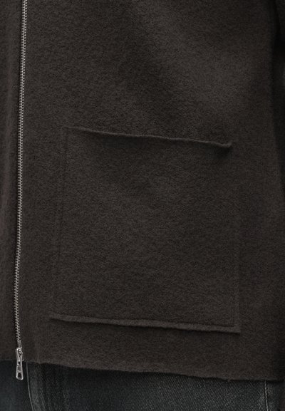 Brązowa wełniana kurtka o teksturowanym wykończeniu, z srebrnym zamkiem błyskawicznym i dużą, kwadratową naszywaną kieszenią z przodu.