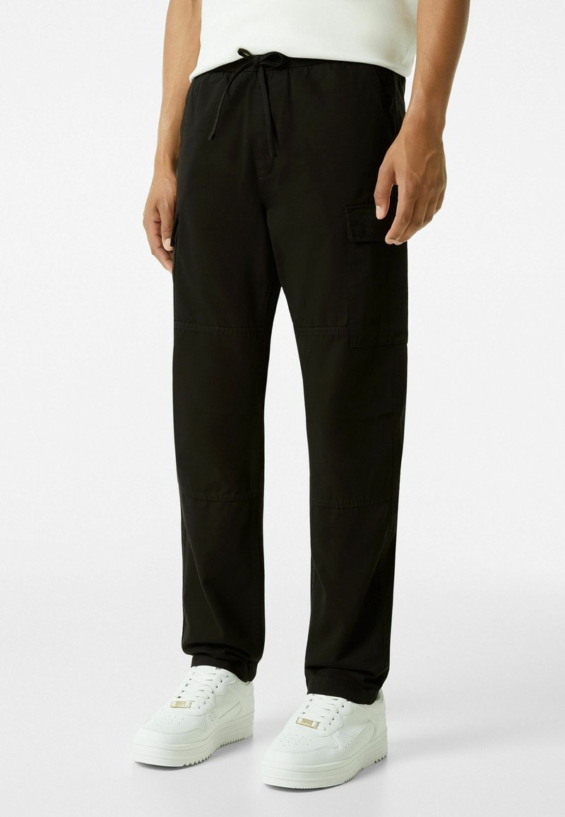 Svarta, lätta byxor med elastisk midja har en dragsko, sidfickor och en avslappnad passform. Kombinationen med vita sneakers.