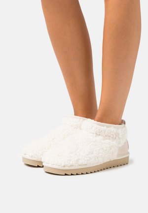 Zapatillas blancas y esponjosas con una textura suave, que presentan un diseño de perfil bajo y una suela beige, mostrando un estilo sencillo y cómodo.
