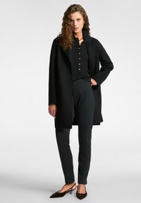 Czarny wełniany płaszcz z wyłami, w połączeniu z czarną koszulą na guziki i ciemnymi spodniami. Brązowe teksturowane buty na obcasie dopełniają look.