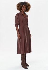 Brązowa sukienka kopertowa z długimi rękawami, zapięciem na guziki i plisami, zestawiona z brązowymi zamszowymi kozakami do kolan. Gładka faktura materiału.