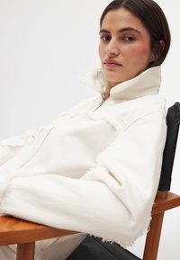 Valge tõmbluku kaelusega jakk kulunud servadega, valmistatud pehmest kangast. Mudel istub, olles riietatud jakki, millel on kõrge kaelus ja lõdva istuvusega. 