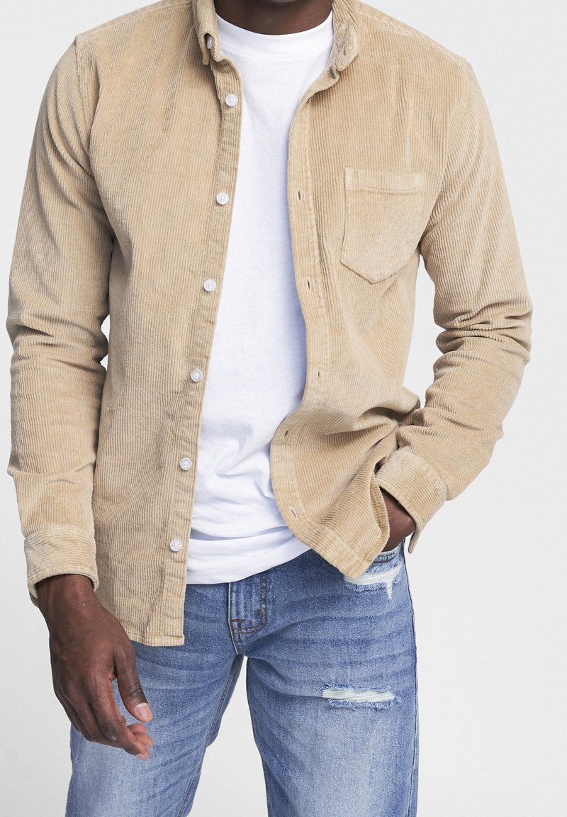 Chemise boutonnée en velours côtelé beige avec une poche poitrine, portée sur un t-shirt blanc ; associée à un jean déchiré bleu clair.