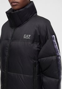 Άτομο φοράει μαύρο μπουφάν puffer με ψηλό γιακά και λογότυπο EA7 Emporio Armani στο στήθος και στη ρίγα του μανικιού.