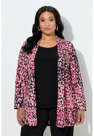 Žena s dlouhými černými vlasy, která má na sobě velké kruhové náušnice, dlouhý růžovo-černý vzorovaný kabát přes černý top a kalhoty, stojící na bílém pozadí.