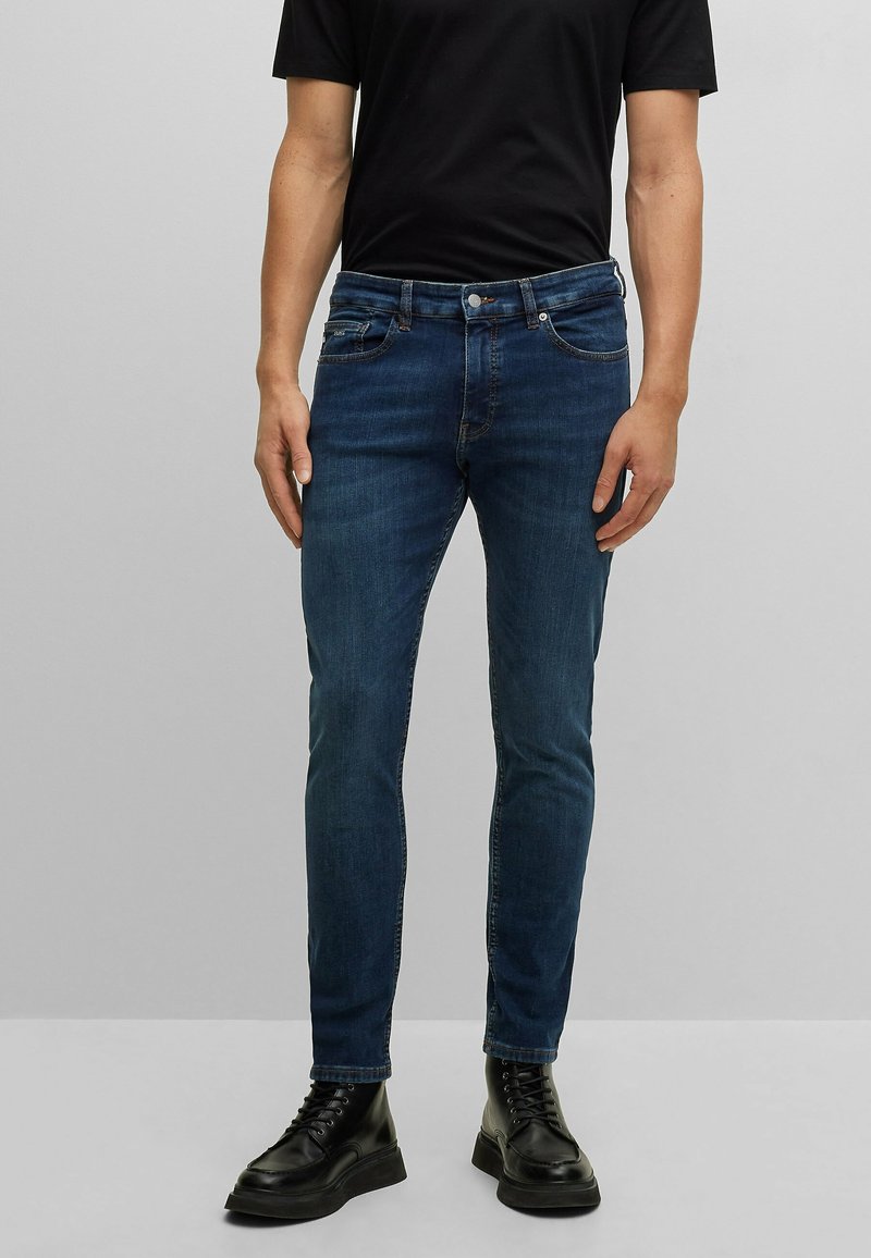 Hombre que lleva jeans oscuros ajustados, camiseta negra y botas negras con cordones, contra un fondo gris claro liso.