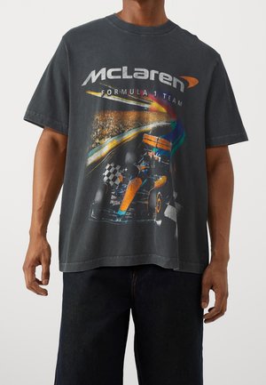 Maglietta nera con una vettura da corsa McLaren di Formula 1 che taglia il traguardo su una pista, indossata da una persona con pantaloni scuri.