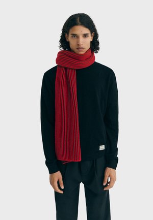 Rote und schwarze gerippte Strickschal, der über einem schwarzen Langarmshirt getragen wird, mit einem strukturierten Design und einem Etikett am Saum.