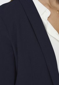 Granatowa marynarka o gładkiej fakturze i wyraźnych klapach, noszona na białej koszuli. Charakteryzuje się prostymi liniami i dopasowanym krojem.