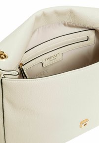 Interno della borsa bianca con texture, dotato di dettagli in metallo dorato e un'etichetta con il marchio. Include una tasca con zip e foderato in modo liscio.