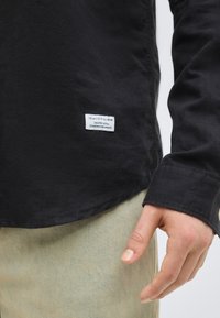 Μαύρο μπλουζάκι με μακριά μανίκια και μια μικρή λευκή ετικέτα κοντά στη βάση, φορεμένο με ανοιχτό μπεζ παντελόνι, με το δεξί χέρι χαλαρό δίπλα στο σώμα.