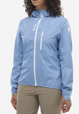 Femme portant une veste de pluie Millet bleu clair avec des fermetures éclair blanches et un pantalon beige, montrée du cou aux hanches.