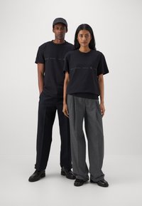 Camisetas de algodón negras con logotipos bordados, combinadas con pantalones oscuros y pantalones anchos grises. Ambas figuras llevan calzado negro.
