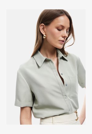 Kvinde med langt brunt hår har en lysegrå kortærmet skjorte med knapper, brystlomme og store guld hoop-øreringe.