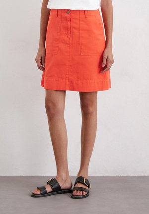 Jupe en coton orange avec deux poches avant, fermeture à bouton et coupe droite. Assortie à des sandales noires avec une boucle en or.