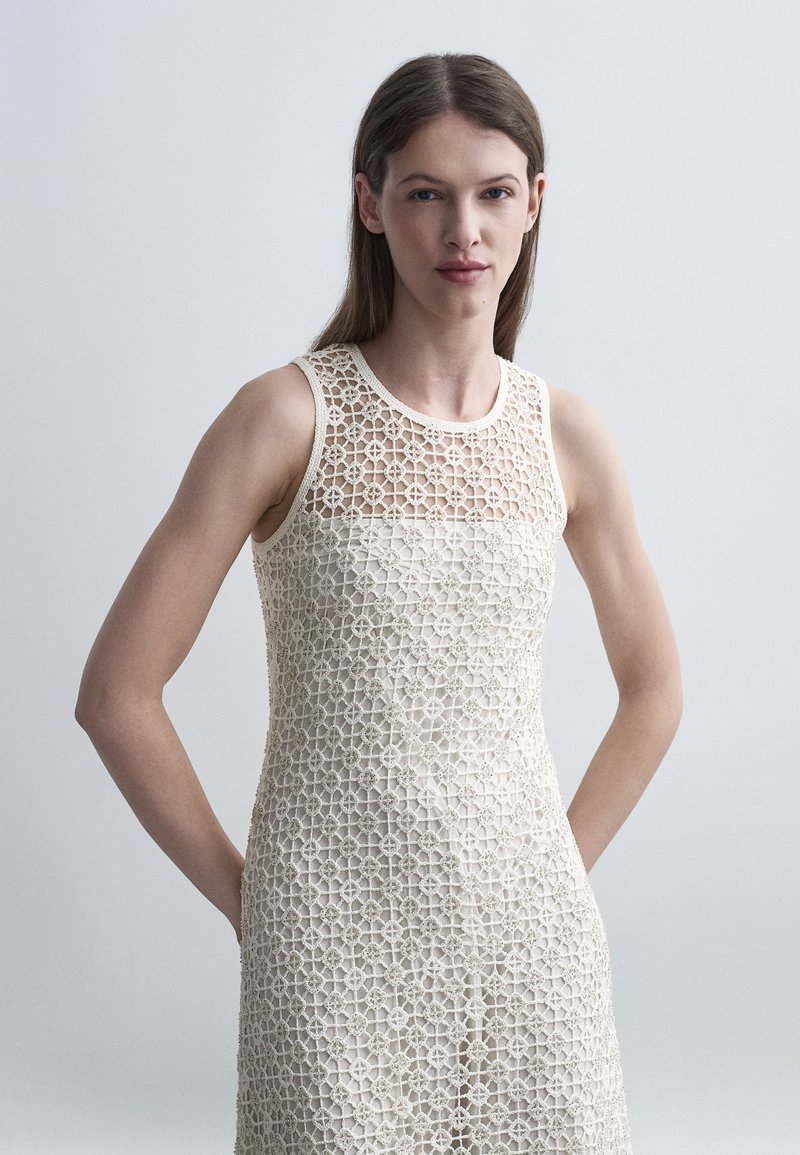 Femme aux longs cheveux bruns portant une robe en crochet beige sans manches avec motif floral, debout devant un arrière-plan gris clair uni.