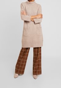 Abito lavorato a maglia beige con colletto alto, due tasche frontali e maniche a tre quarti, abbinato a pantaloni a gamba larga a quadri marroni e tacchi beige.