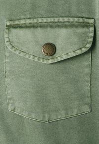 Hellgrüner Denim-Stoff, verziert mit einer Klapptasche. Die Tasche ist mit einem runden bronzefarbenen Knopf gesichert, auf dem der Markenname eingraviert ist.