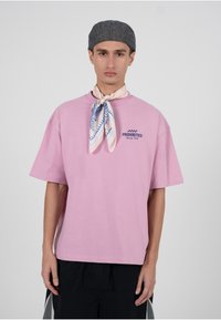 Roosa puuvillane lühikeste varrukatega T-särk, millel on tumesasine "PROHIBITED Racing Club" logo. Kantud koos mustrilise salliga ja halli nokamütsiga.