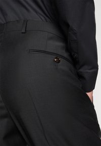 Pantalons noirs ajustés, dotés d'une texture lisse, d'une poche arrière avec fermeture à bouton, et de détails de couture subtils.