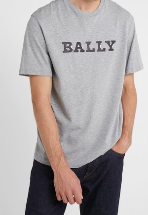 Mężczyzna w szarej koszulce z krótkim rękawem z dużym napisem „BALLY” w zestawieniu z ciemnymi dżinsami.