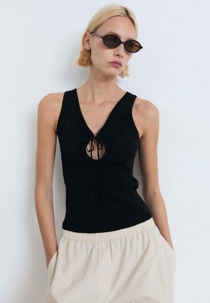 Schwarzes Strick-Top mit tiefem V-Ausschnitt und einem Ausschnitt-Detail mit Knotenverschluss. Kombiniert mit hellen, lässig sitzenden Shorts.
