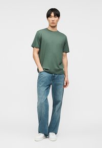 Νέος άνδρας που φοράει πράσινο t-shirt με κοντά μανίκια, φαρδιά μπλε τζιν και λευκά αθλητικά παπούτσια, στέκεται με το ένα χέρι στην τσέπη του μπροστά σε απλό φόντο.