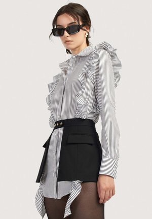 Naine mustade päikeseprillidega, triibulise volangiga pluusi, musta asümmeetrilise miniseeliku ja läbipaistvate mustade sukkadega, seisab külje pealt ühevärvilise tausta ees.