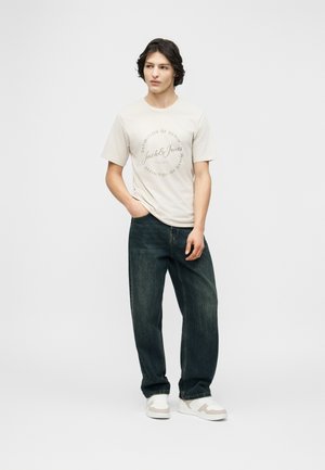 Jonge man die een beige Jack & Jones T-shirt draagt, donkere losse spijkerbroek en witte sneakers, staand met één hand in de zak tegen een effen achtergrond.