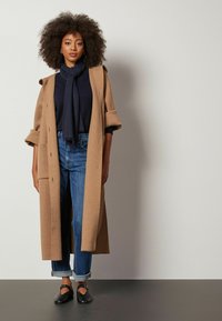 Lang kamelfrakke over en marineblå sweater, parret med opkørselskanter i blå jeans og sorte flade sko, med en afslappet pasform og minimalistisk design.