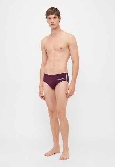 Junger Mann, der barfuß steht und maroonfarbene Badehosen mit weißen Seitenstreifen und dem DSQUARED2-Logo auf einem schlichten weißen Hintergrund trägt.