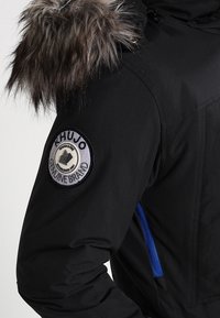 Veste d'hiver noire avec capuche bordée de fourrure, poche zippée bleue et patch circulaire « Khujo Genuine Brand » sur le haut de la manche.