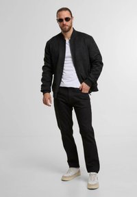 Zwart suède bomberjack met een rits sluiting, gecombineerd met een wit T-shirt en zwarte broeken, aangevuld met beige en witte sneakers.
